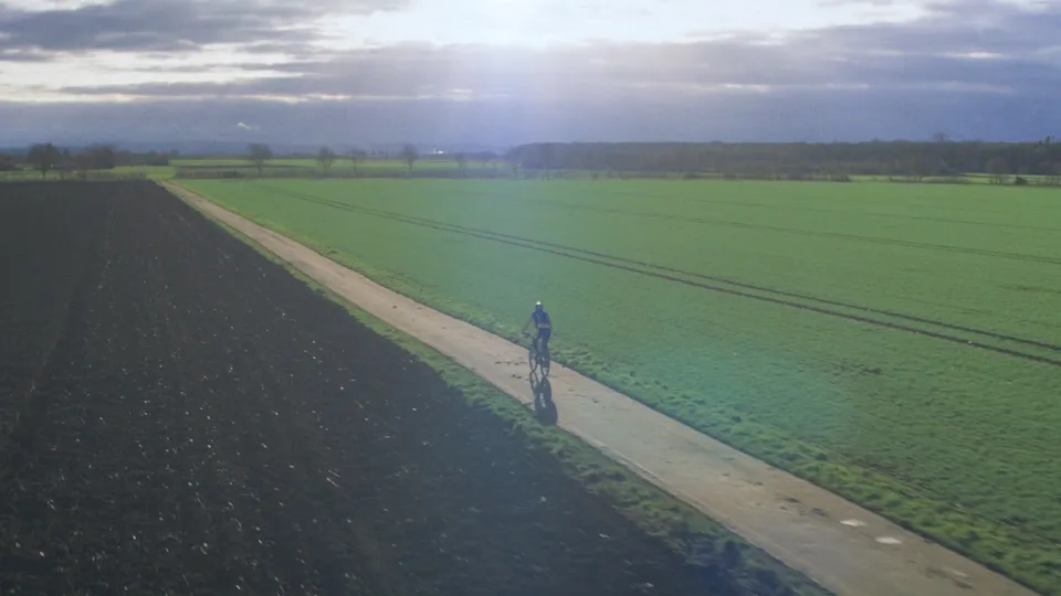 Luftaufnahme eines Radfahrers auf einem Feldweg zwischen dunklem Ackerboden und grüner Wiese unter bewölktem Himmel.