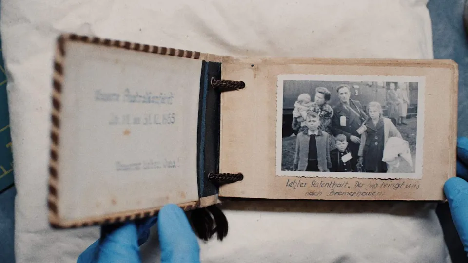 Behandschuhte Hand hält ein geöffnetes altes Fotoalbum mit einem schwarz-weißen Familienfoto und handschriftlicher Bildunterschrift auf vergilbten Seiten.