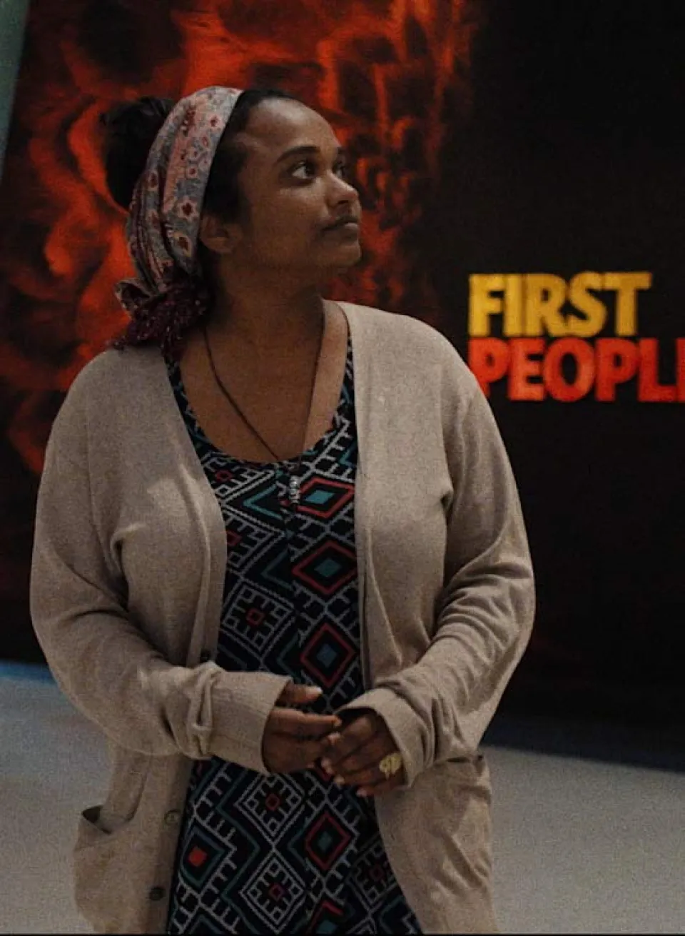Frau mit gemustertem Kleid und beiger Strickjacke steht vor einer Ausstellungswand mit der Aufschrift "FIRST PEOPLE"