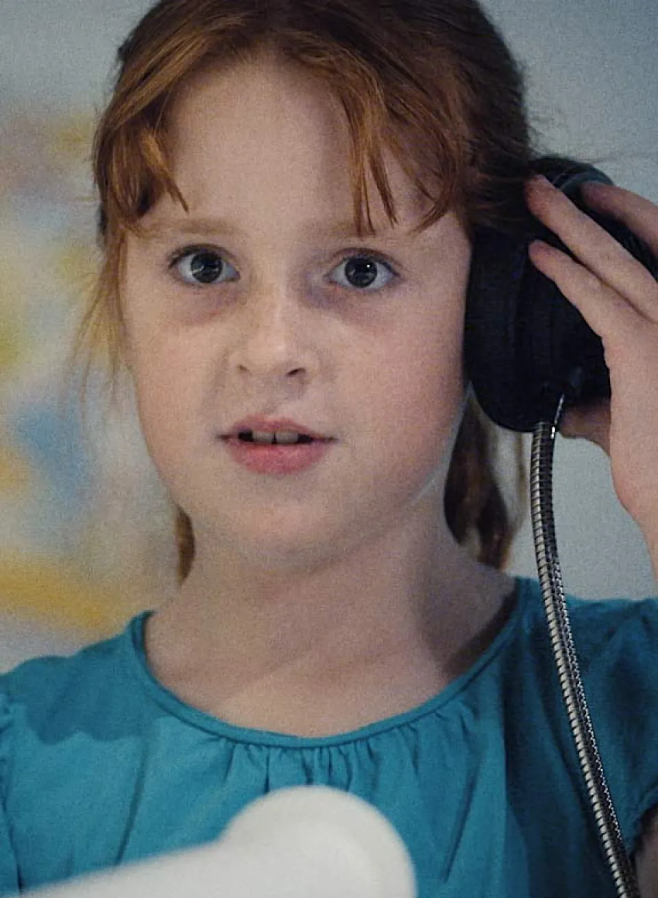 Kind mit rötlich-blondem Haar hält einen schwarzen Telefonhörer ans Ohr und trägt ein türkisfarbenes Oberteil.
