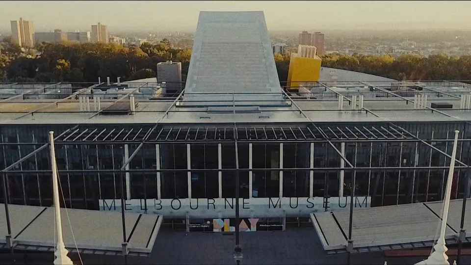 Luftaufnahme des Melbourne Museums mit seiner charakteristischen grauen Dachkonstruktion, gläserner Fassade mit Schriftzug und Stadtpanorama im Hintergrund.