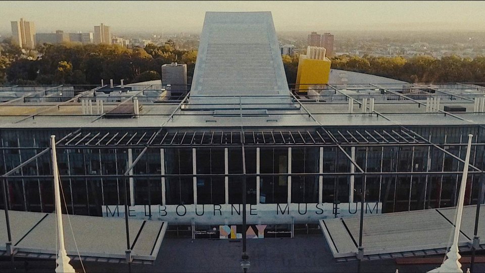Luftaufnahme des Melbourne Museums mit seiner charakteristischen grauen Dachkonstruktion, gläserner Fassade mit Schriftzug und Stadtpanorama im Hintergrund.