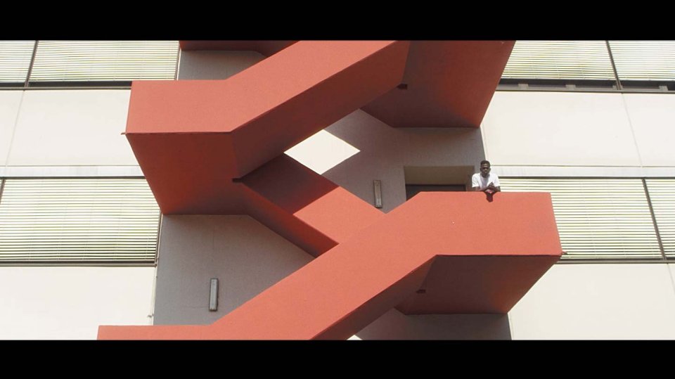 Person steht auf korallenroter Außentreppe an moderner Gebäudefassade mit horizontalen Jalousien, Ansicht von unten.