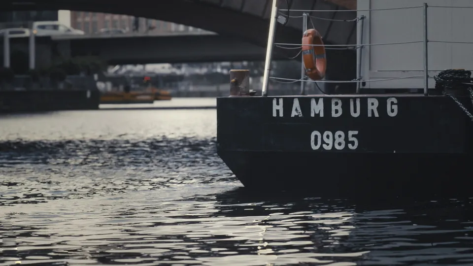 Schiffsbug mit Aufschrift "HAMBURG 0985" und orangefarbenem Rettungsring an der Reling im Hafenwasser