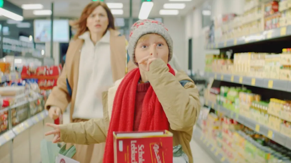 Kind in beiger Winterjacke und roter Weste isst im Supermarkt, während eine Frau im Hintergrund mit Einkaufswagen folgt.