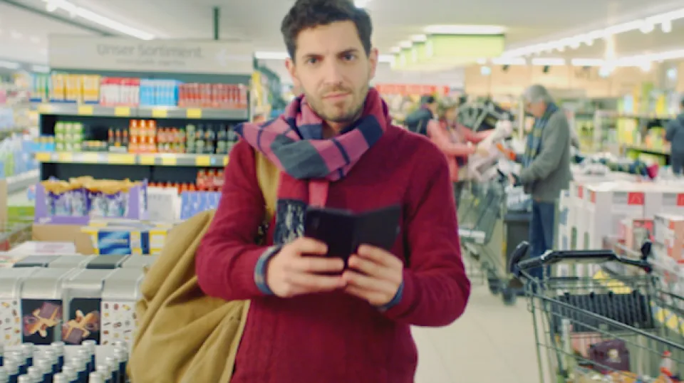 Mann mit Smartphone und Schal steht in einem Supermarkt, im Hintergrund Regale mit Produkten und weitere Kunden.