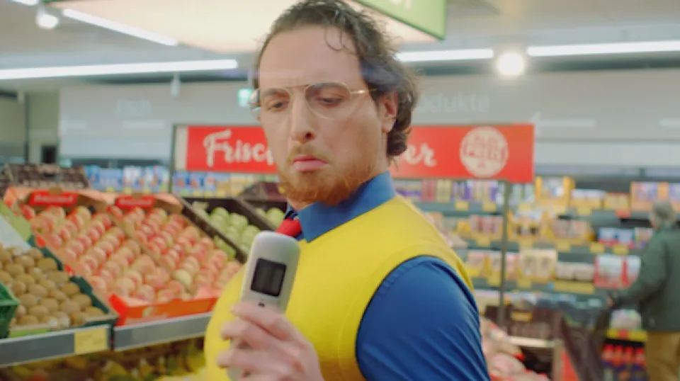 Mann mit Brille und rot-gelbem Bart in blau-gelber Arbeitskleidung hält Barcode-Scanner in einem Supermarkt vor der Obst- und Gemüseabteilung.