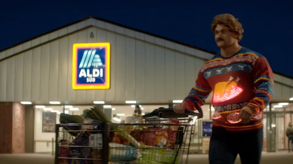 Mann in weihnachtlichem Pullover mit leuchtendem Herz steht mit Einkaufswagen vor einem ALDI SÜD Supermarkt bei Nacht.