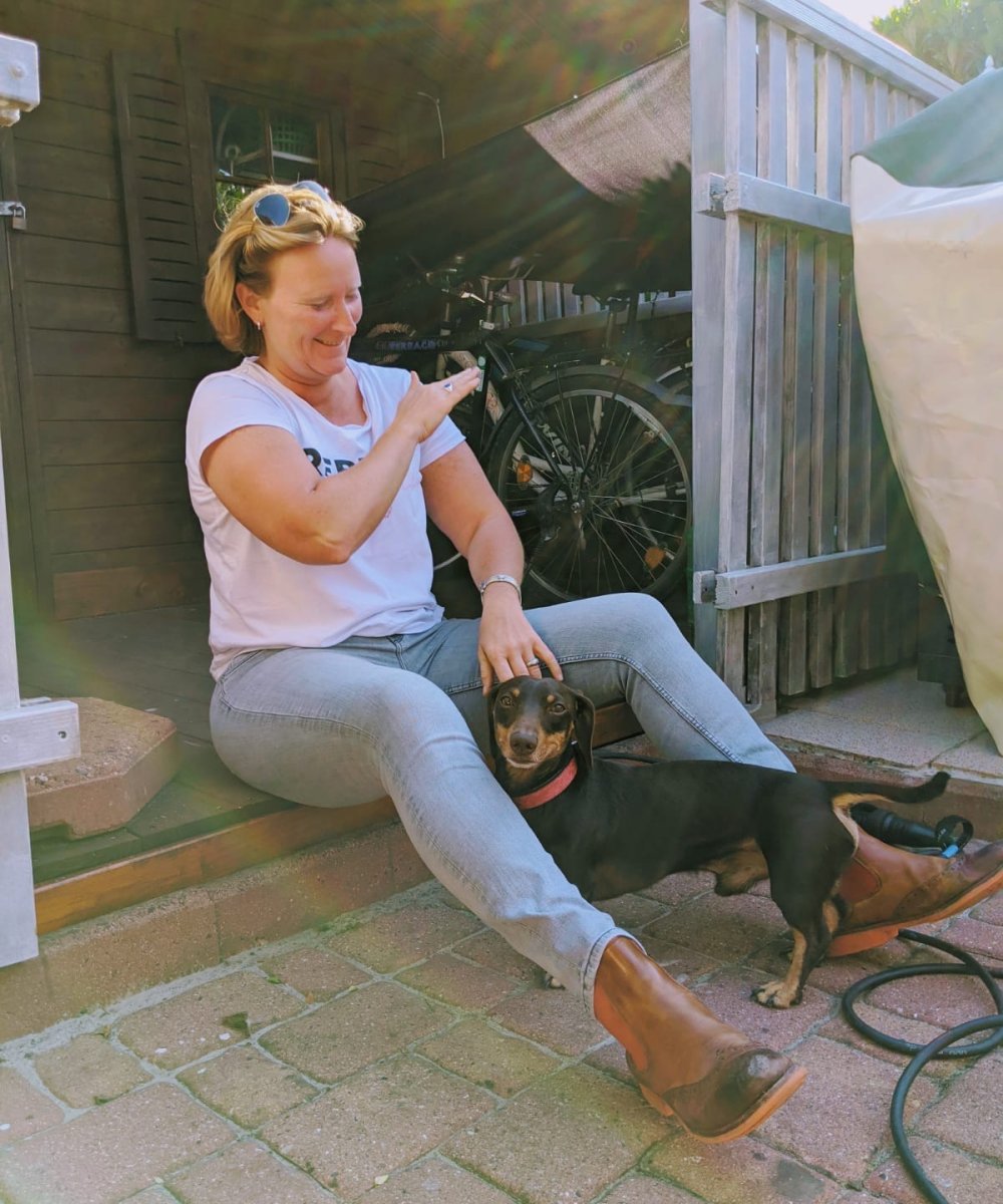 Frau mit Sonnenbrille auf dem Kopf sitzt auf Terrassenstufe und streichelt einen schwarz-braunen Dackel mit rotem Halsband.