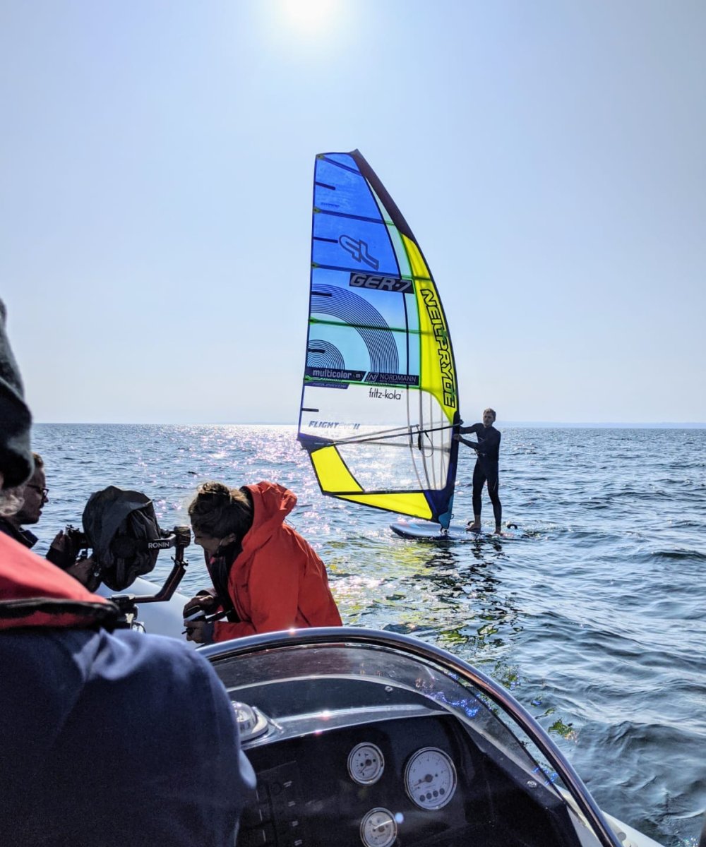 Windsurfer mit blau-gelbem Segel auf ruhigem Meer, fotografiert von einem Boot mit Zuschauern im Vordergrund bei Sonnenschein.