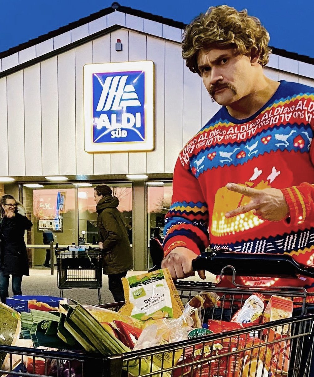 Mann mit blonder Perücke und buntem ALDI-Pullover steht mit vollem Einkaufswagen vor einem ALDI Süd Supermarkt in der Abenddämmerung.