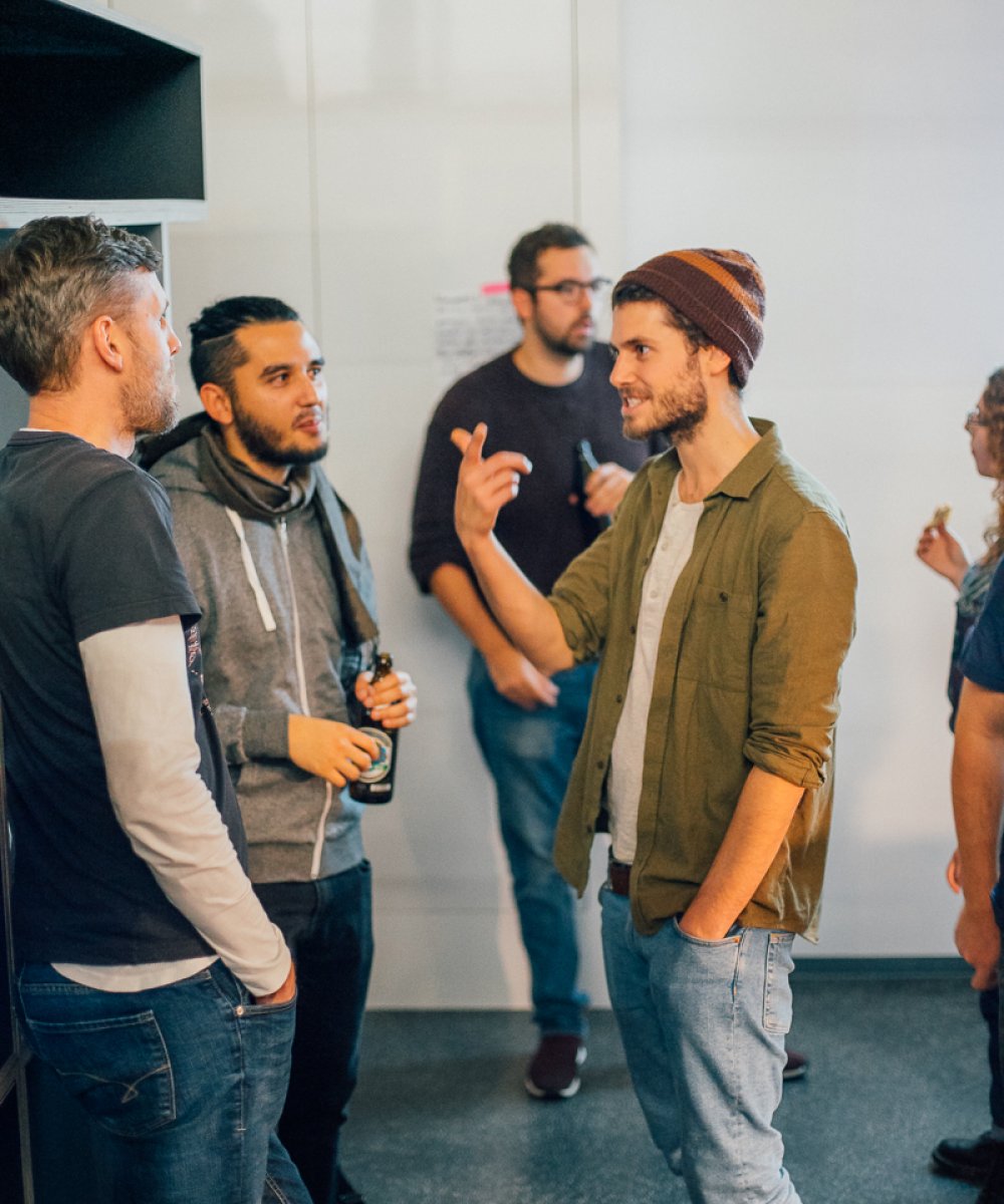 Fünf junge Männer stehen in lockerem Kreis in einem hellen Büroraum und führen eine lebhafte Diskussion, ein Mann gestikuliert dabei mit den Händen.