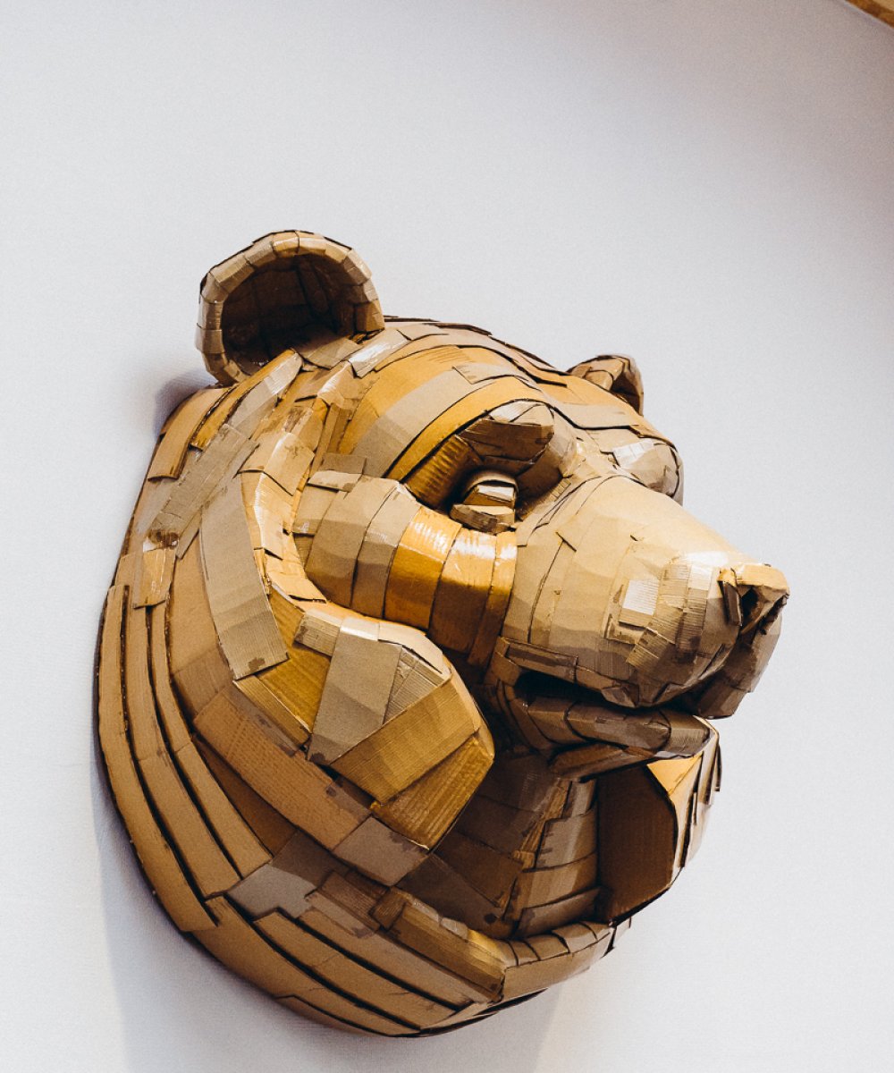 Dreidimensionale Wandskulptur eines Bärenkopfes aus geschichtetem Holz in natürlichen Brauntönen an weißer Wand.