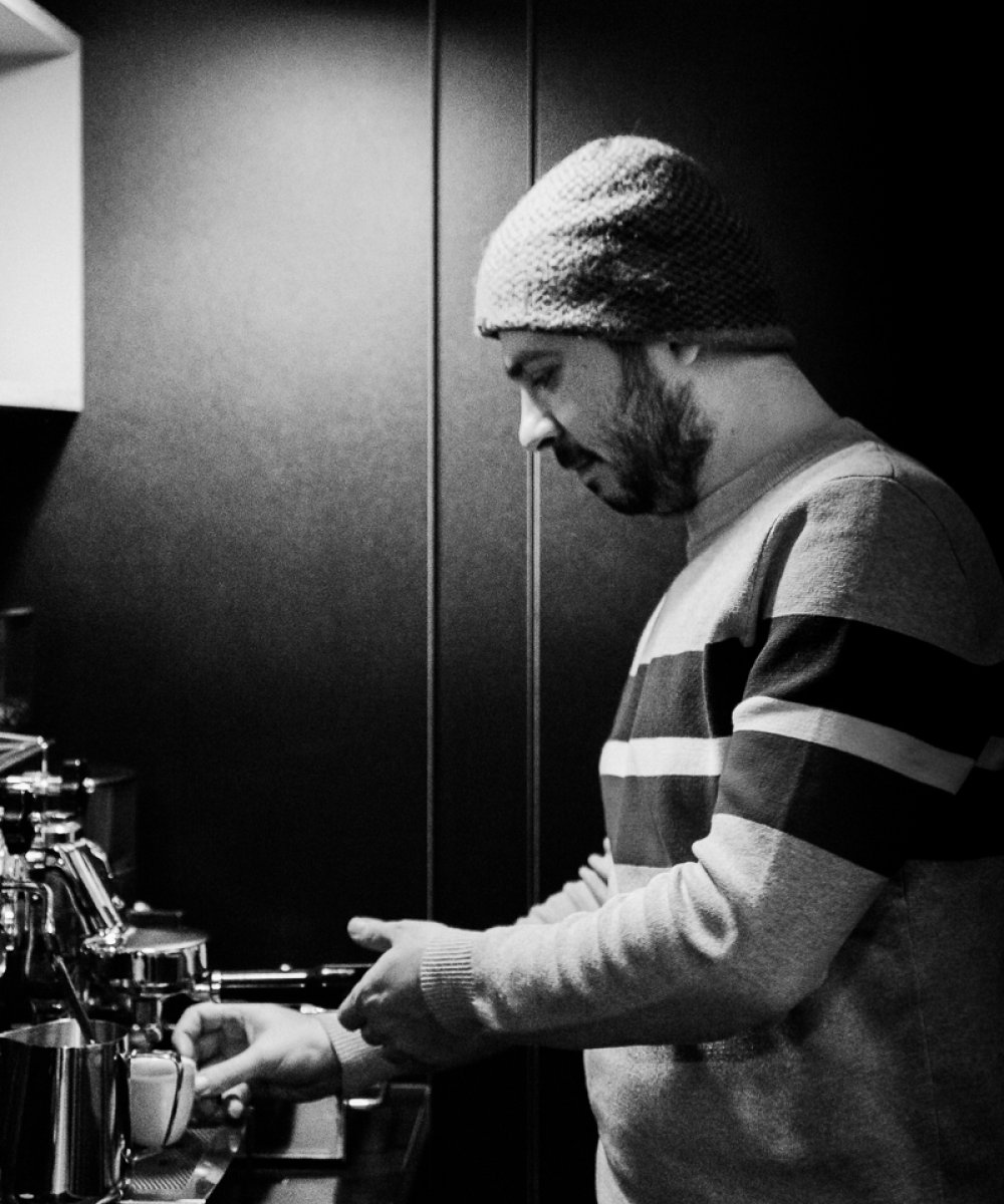 Mann mit Strickmütze bereitet Kaffee an einer Espressomaschine zu, Schwarzweißfotografie mit Deckenbeleuchtung.