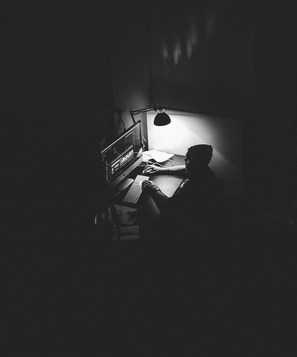 Person arbeitet spät nachts am beleuchteten Schreibtisch mit Computer in dunklem Raum, Schwarz-Weiß-Fotografie.