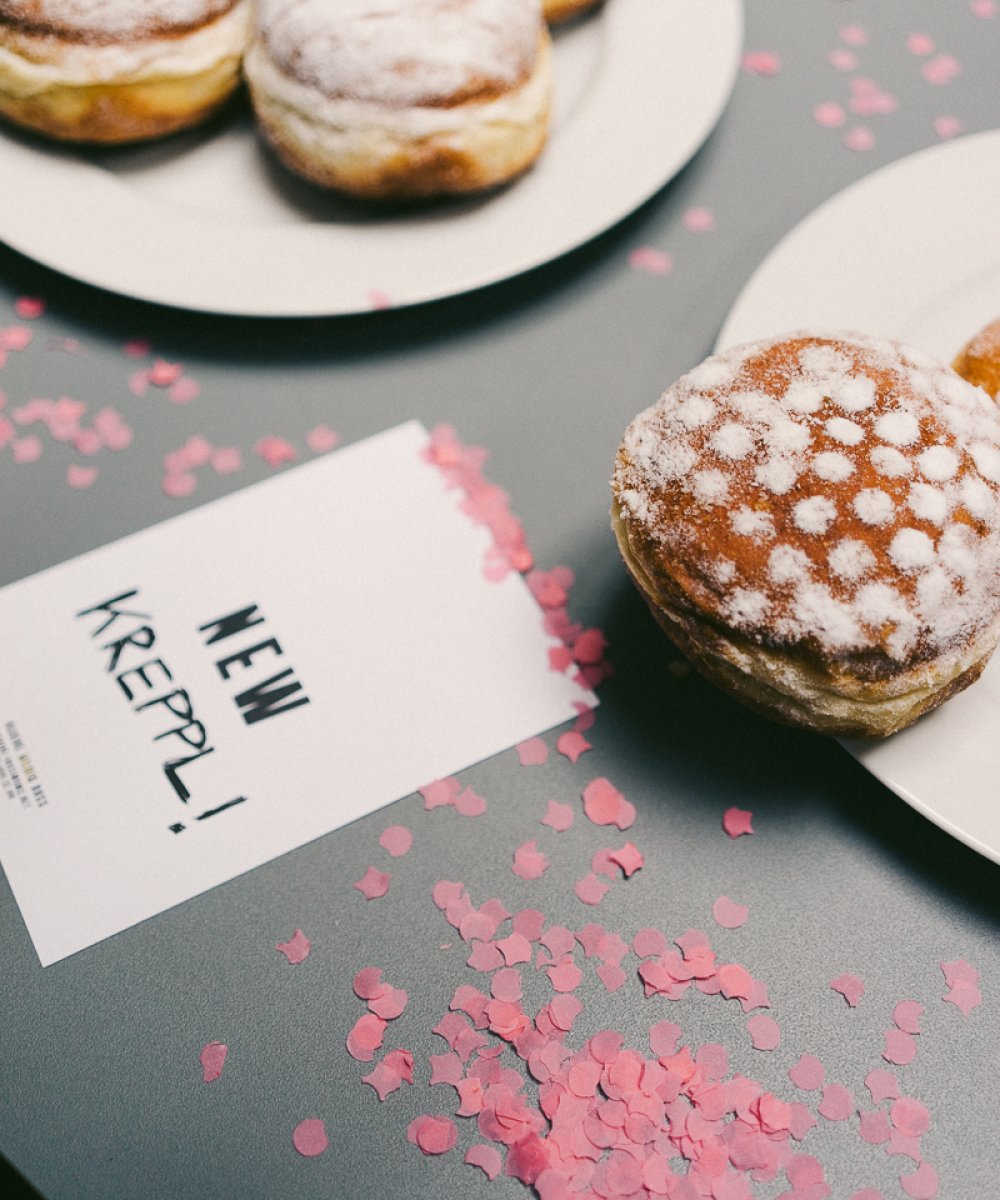 Mit Puderzucker bestäubte Krapfen auf weißen Tellern, daneben eine Karte mit der Aufschrift "NEW KREPPL!" und rosa Konfetti auf grauer Oberfläche.