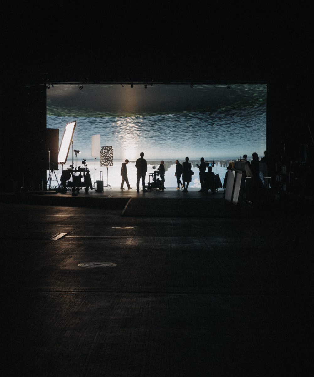 Filmset in dunkler Halle mit Crew-Silhouetten vor hell beleuchtetem Green Screen, der einen Ozean mit Wasseroberfläche zeigt.