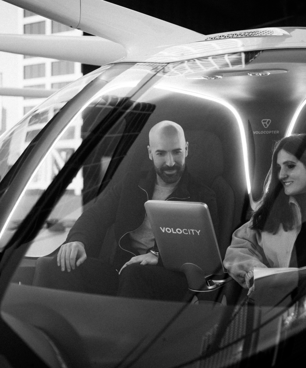 Zwei Personen sitzen im Cockpit eines Volocopter-Flugtaxis, ein Mann am Steuer und eine Frau als Passagierin, während eine Person im Hintergrund steht.