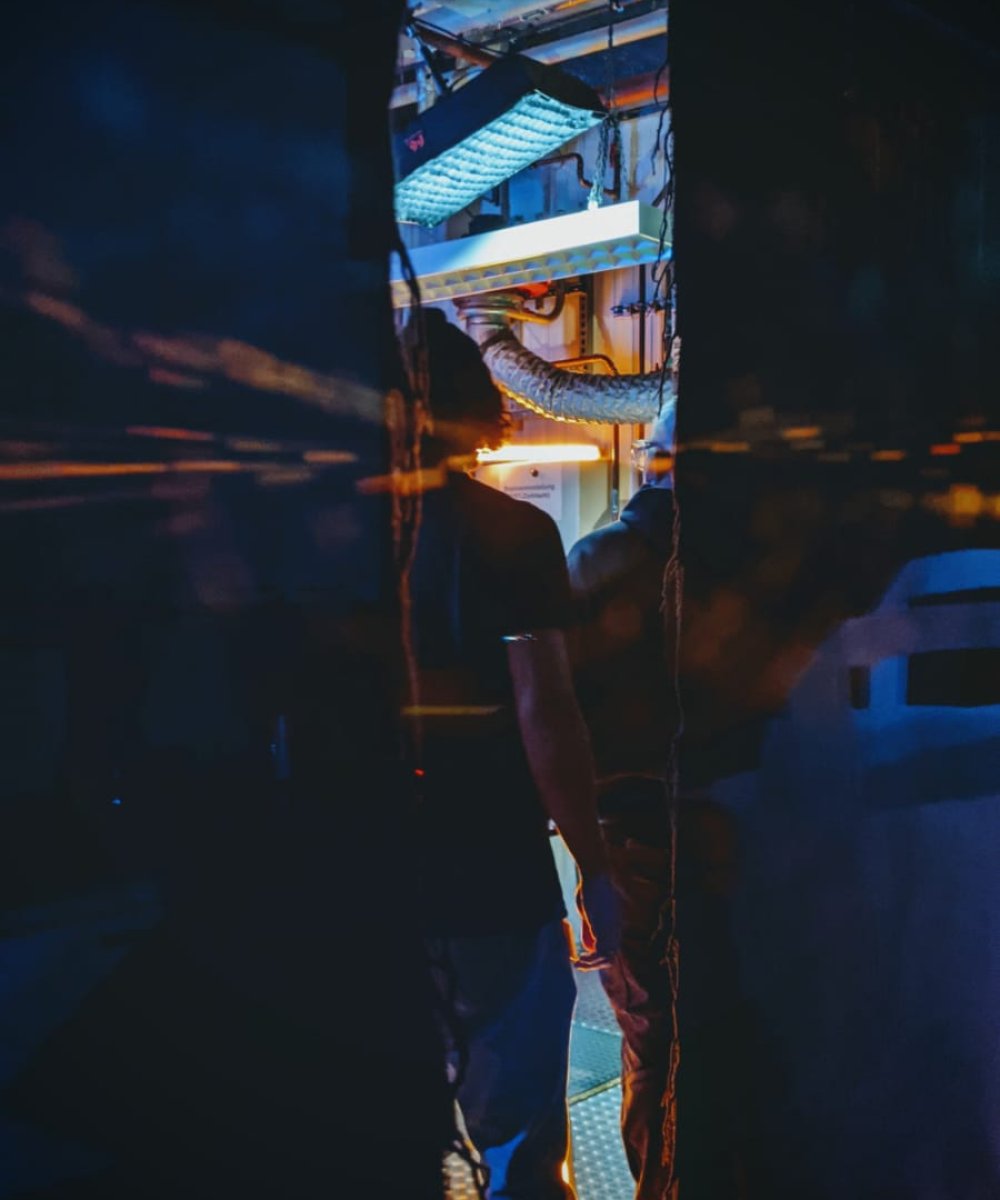 Spiegelung eines beleuchteten mehrstöckigen Gebäudes in einer Wasserpfütze bei Nacht, mit Silhouetten von Personen im Vordergrund.