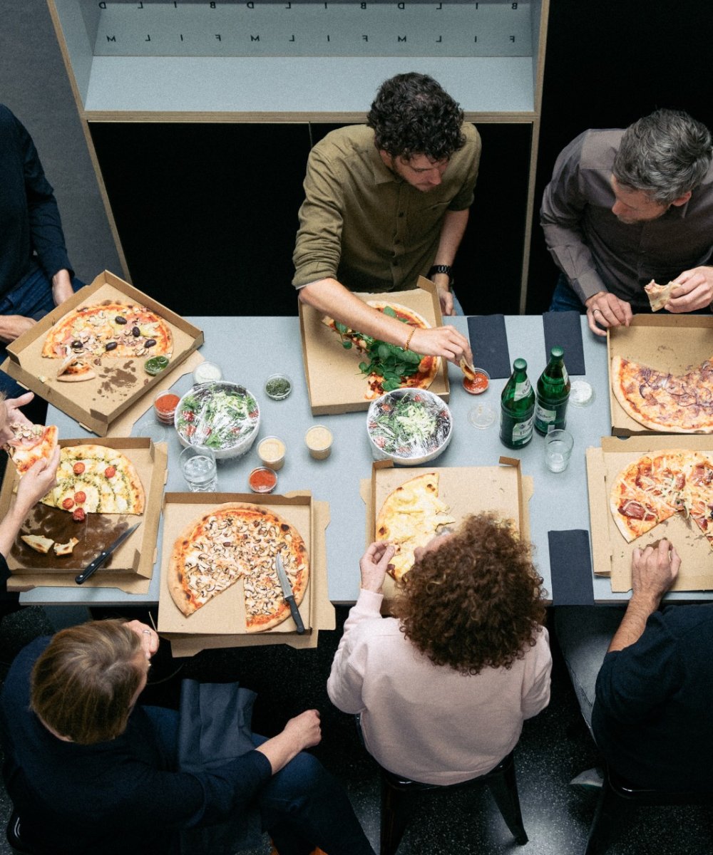 Vogelperspektive einer Gruppe von sieben Personen, die gemeinsam Pizza aus Kartons an einem Tisch in einem Büro essen.