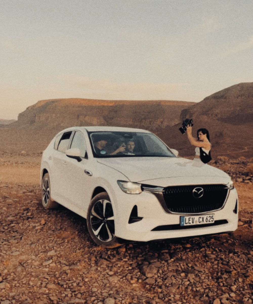 Weißer Mazda CX-625 SUV in steiniger Wüstenlandschaft bei Sonnenuntergang, Frau fotografiert aus dem Seitenfenster, Felsklippen im Hintergrund