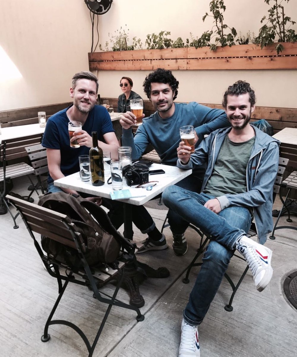 Vier Freunde sitzen an einem Tisch in einem Café mit Biergläsern, im Hintergrund eine Person mit Sonnenbrille und Pflanzen auf einem Holzregal.