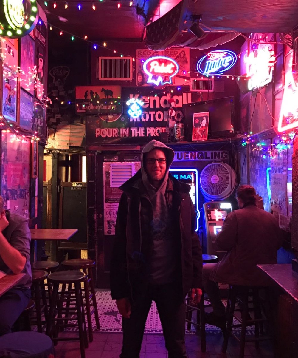 Mann mit Hut und Brille steht in einer bunt beleuchteten Bar mit Neonschildern, Lichterketten und nostalgischer Dekoration an den Wänden.