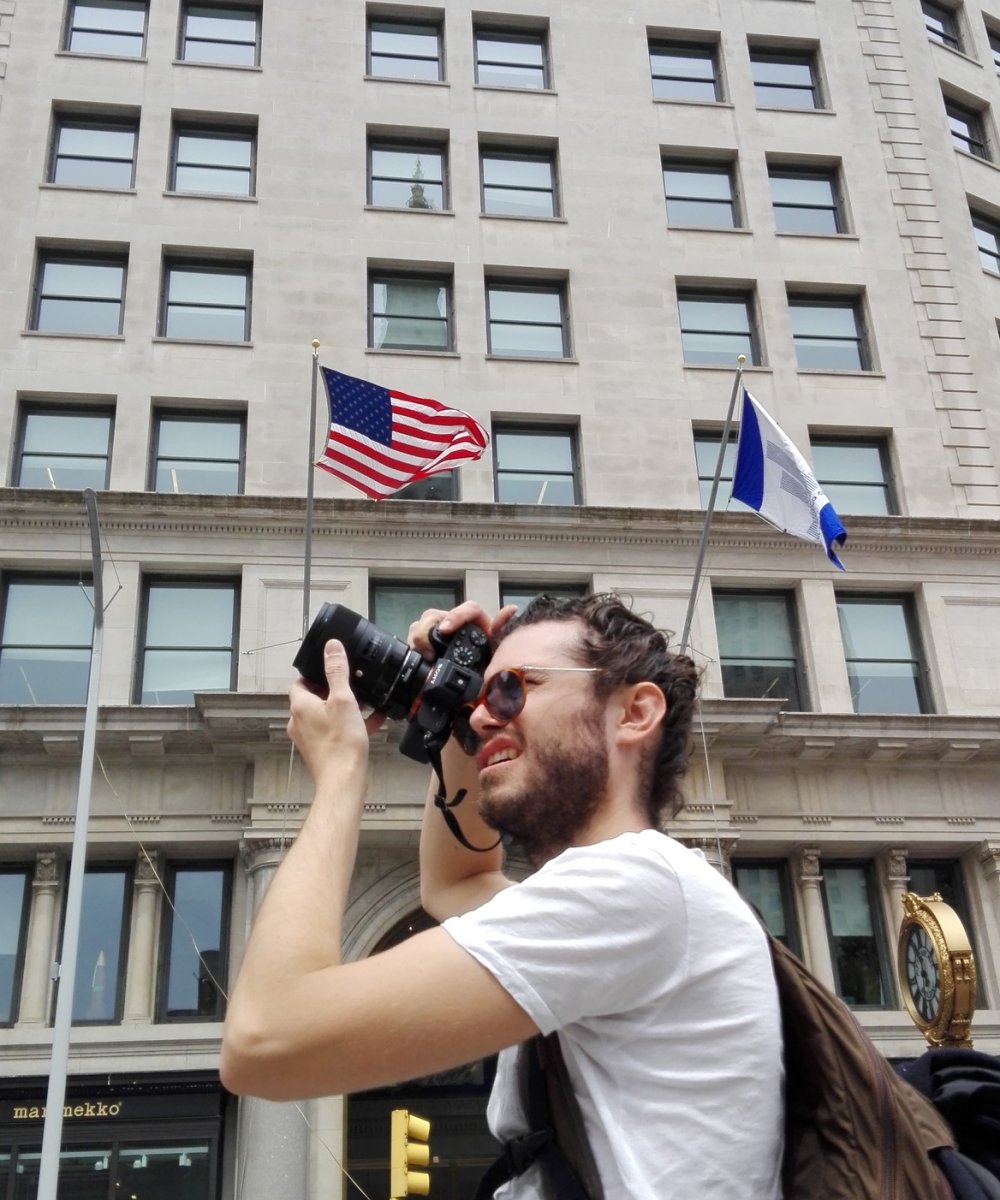 Mann mit Sonnenbrille fotografiert mit Kamera vor einem mehrstöckigen Gebäude mit amerikanischer Flagge.