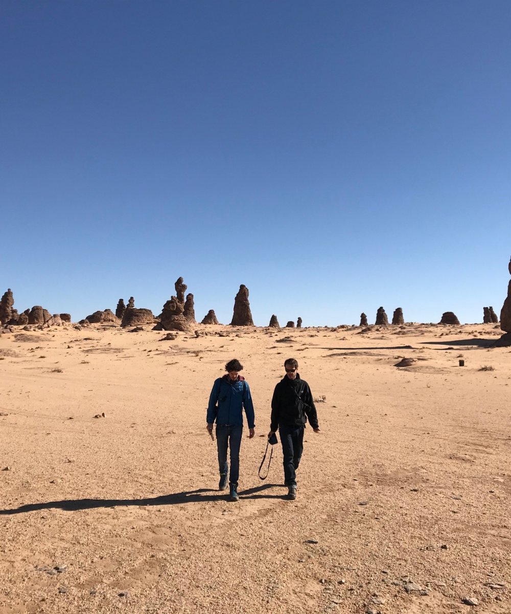 Zwei Personen wandern durch eine Wüstenlandschaft mit zahlreichen hohen Sandsteinfelsen unter blauem Himmel.