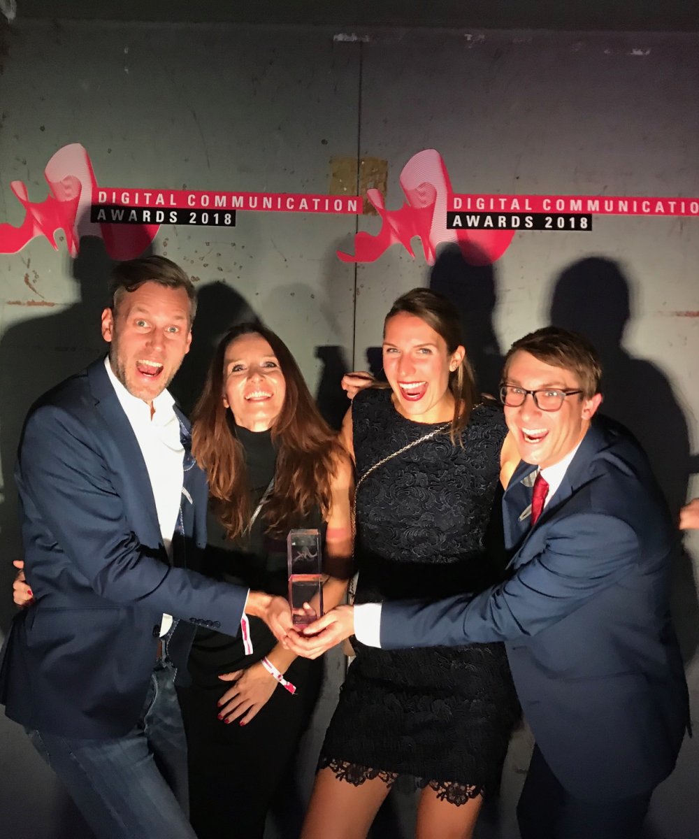 Vier Personen in festlicher Kleidung posieren fröhlich mit einem Award vor einer Wand mit dem Logo "Digital Communication Awards 2018".