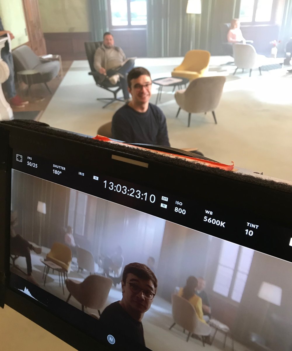 Blick durch einen Kameramonitor auf ein Filmset mit zwei Personen in einem modern eingerichteten Raum mit Sesseln und Fenstern.