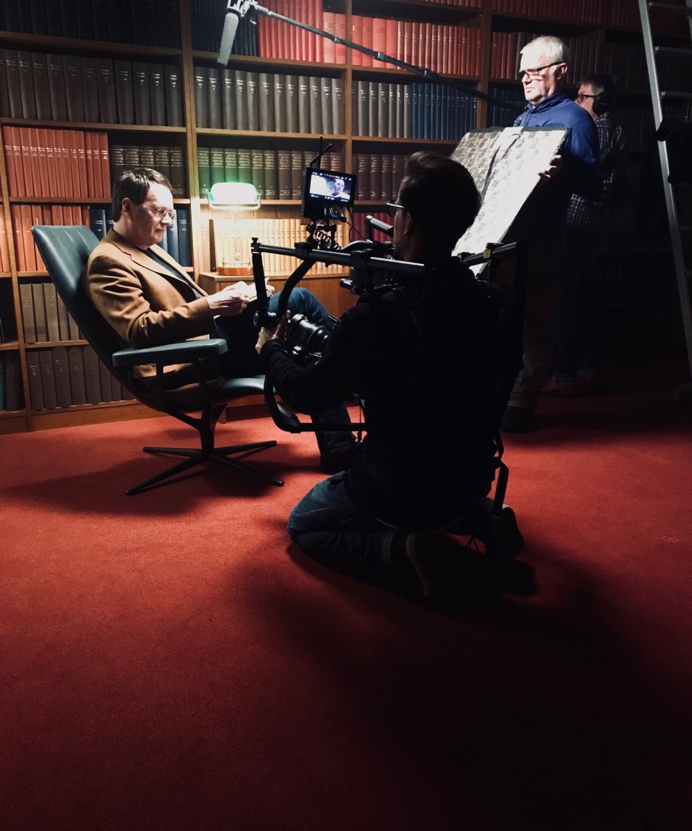 Filmdreh in einer Bibliothek: Mann im braunen Anzug sitzt in grünem Ledersessel, während Kameramann und Crew mit professioneller Ausrüstung und Beleuchtung vor Bücherregalen filmen.