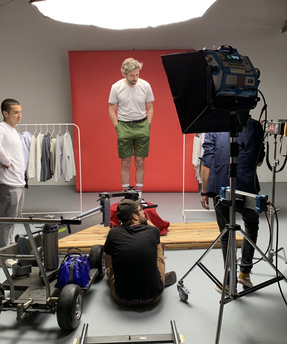 Fotoshooting in einem Studio mit Model vor rotem Hintergrund, Fotograf auf dem Boden sitzend, Kamera-Crew und Beleuchtungsausrüstung, Kleiderständer im Hintergrund.