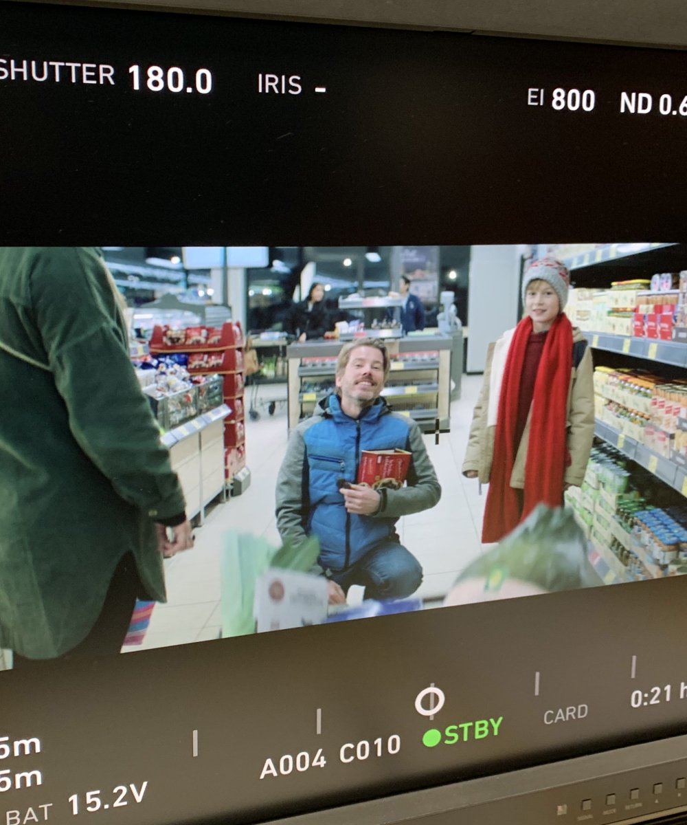 Kameramonitor zeigt Filmaufnahme in einem Supermarkt mit kniender Person in blauer Jacke und zwei Personen in Winterkleidung in den Regalgängen