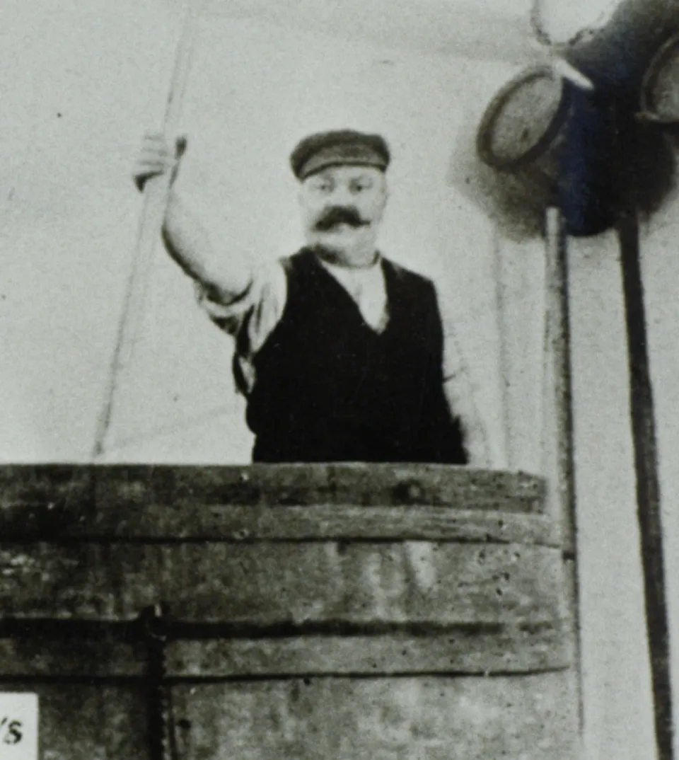 Historisches Schwarzweißfoto eines Mannes mit Schnurrbart und Mütze, der hinter einem Holzzaun steht und grüßend die Hand hebt, mit einer Laterne rechts im Bild.