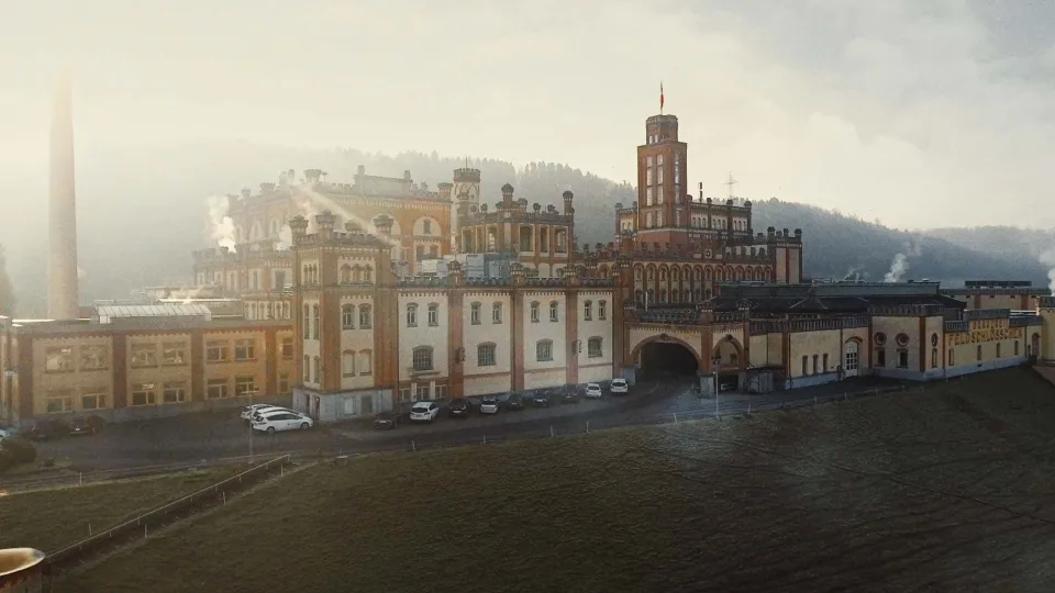 Historisches Industriegebäude mit markanten Türmen und Bögen an einem nebligen Morgen, im Vordergrund parkende Autos, im Hintergrund bewaldete Hügel.