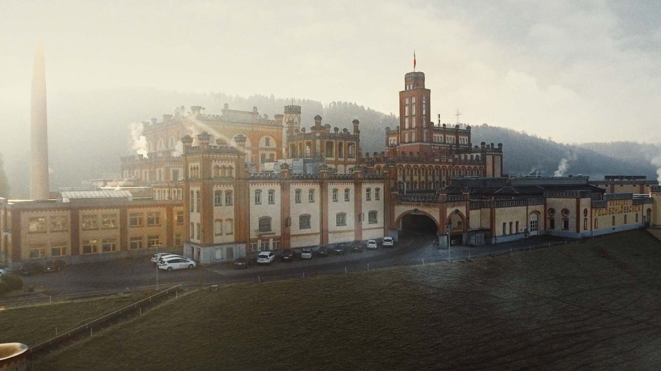 Historisches Industriegebäude mit markanten Türmen und Bögen an einem nebligen Morgen, im Vordergrund parkende Autos, im Hintergrund bewaldete Hügel.