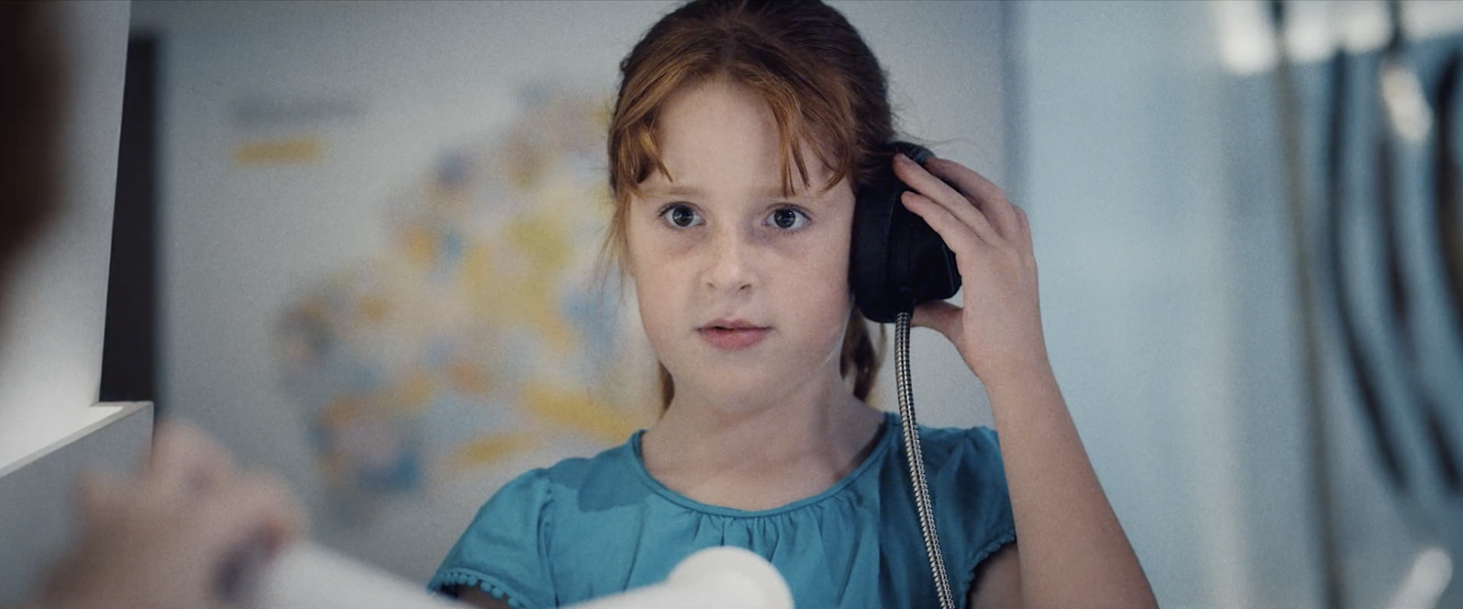 Junges Mädchen in blauem Kleid hält einen schwarzen Telefonhörer ans Ohr und blickt aufmerksam zur Seite.