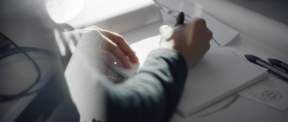Person arbeitet am Schreibtisch mit Computermaus in der linken Hand und Stift in der rechten Hand, umgeben von Dokumenten und Kabeln.