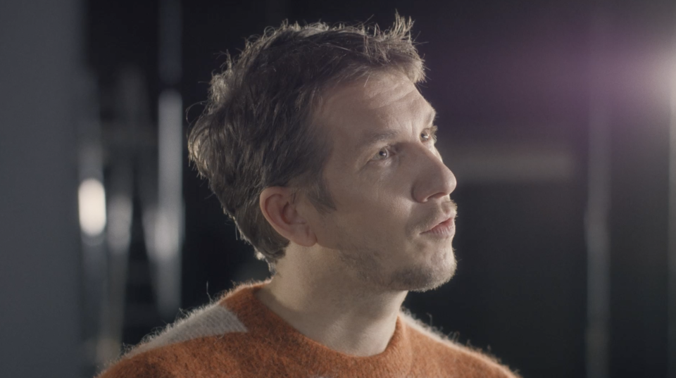 Mann mit kurzem braunem Haar in orangefarbenem Pullover schaut lächelnd nach rechts oben, vor unscharfem dunklem Hintergrund.