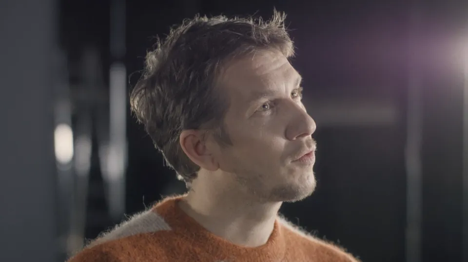 Mann mit kurzem braunem Haar in orangefarbenem Pullover schaut lächelnd nach rechts oben, vor unscharfem dunklem Hintergrund.