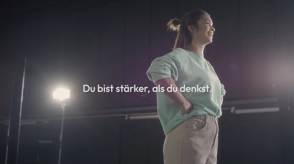 Junge Frau in Sportkleidung steht selbstbewusst lächelnd vor nächtlichem Stadion mit deutscher Motivationstext "Du bist stärker, als du denkst."
