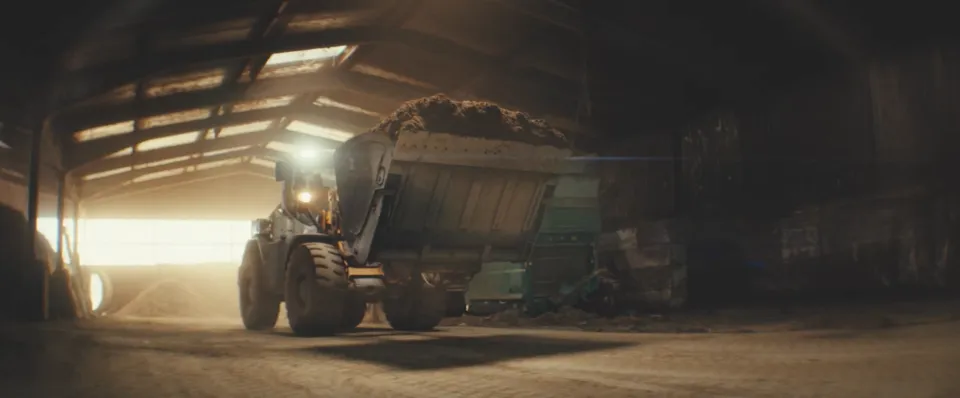 Beladener Traktor mit Anhänger fährt mit eingeschalteten Scheinwerfern durch eine dunkle Scheune ins helle Tageslicht.