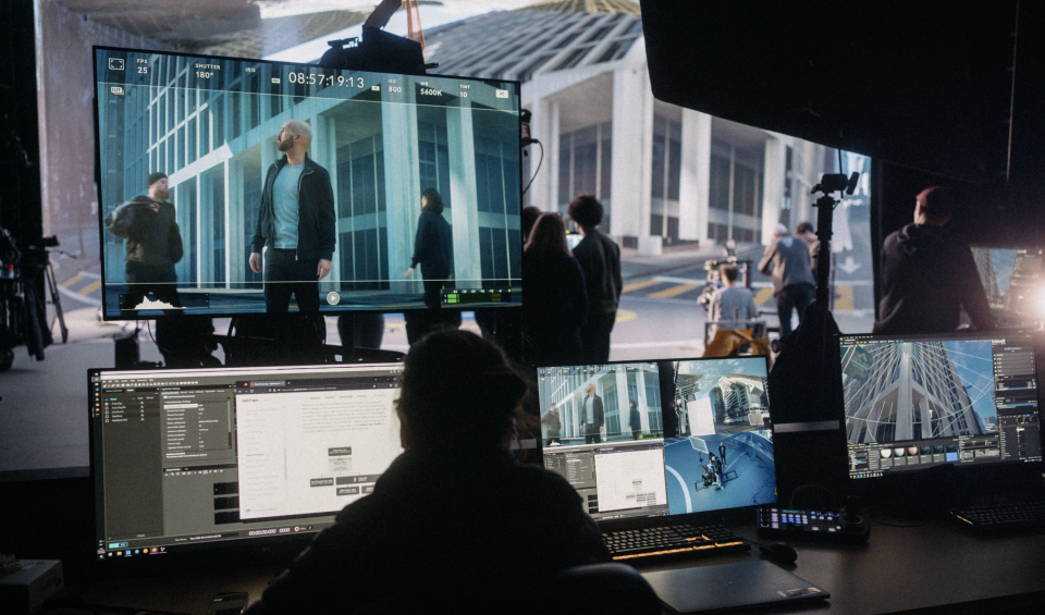 Filmset mit großem Monitor, der eine Szene mit Schauspielern vor einem modernen Gebäude zeigt, während Crew-Mitglieder an mehreren Computern die Aufnahme überwachen.