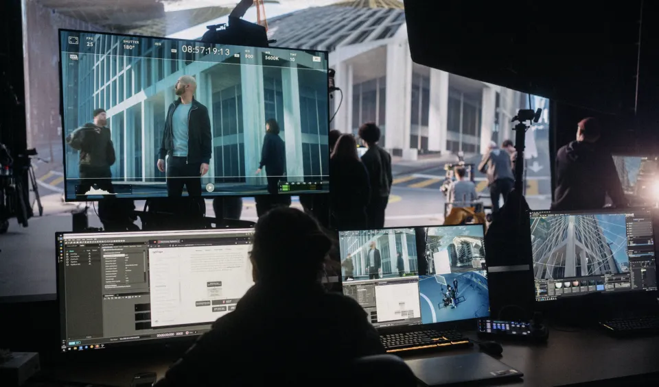 Filmset mit großem Monitor, der eine Szene mit Schauspielern vor einem modernen Gebäude zeigt, während Crew-Mitglieder an mehreren Computern die Aufnahme überwachen.