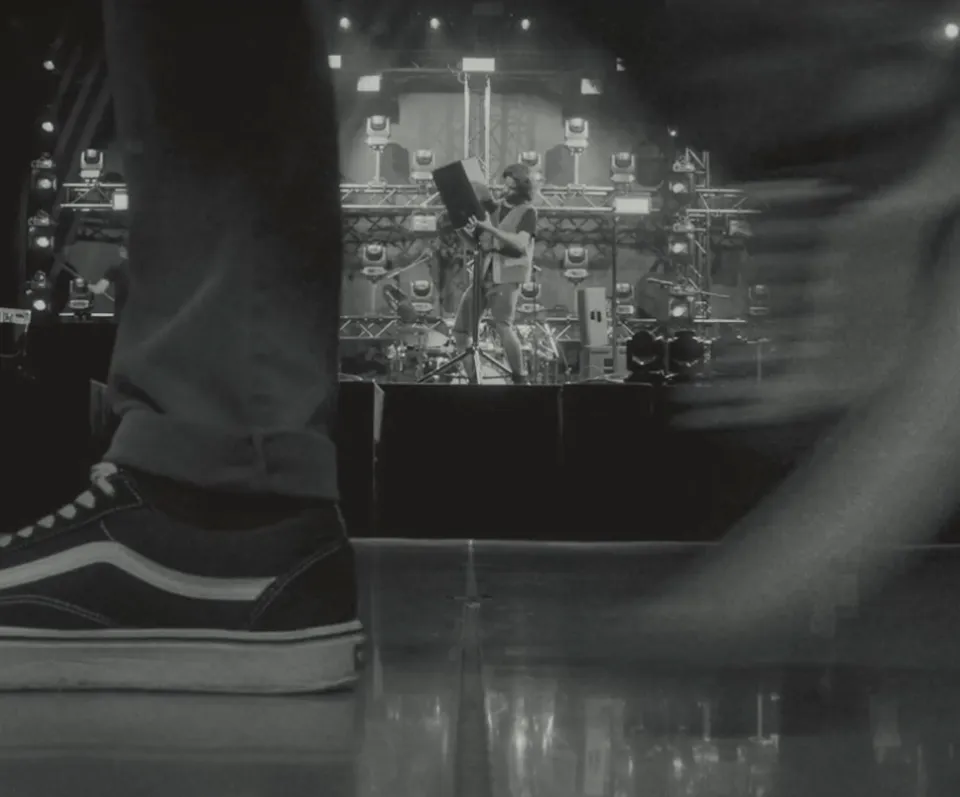 Schwarz-weiß Foto von einem Konzert aus der Perspektive eines Zuschauers, dessen Sneaker im Vordergrund sichtbar ist, während ein Künstler auf der beleuchteten Bühne mit Scheinwerfern und Bühnentechnik performt.