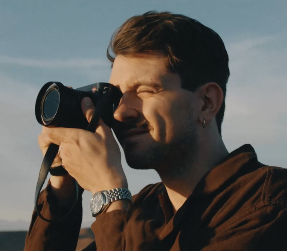 Mann mit Fernglas blickt in die Ferne, trägt Armbanduhr und braune Jacke vor blauem Himmel.