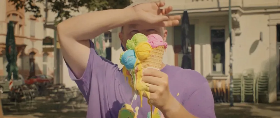 Person in lila T-Shirt hält mehrere schmelzende Eiskugeln in Waffeln vor einem Stadtplatz mit Gebäuden und Bäumen im Hintergrund.