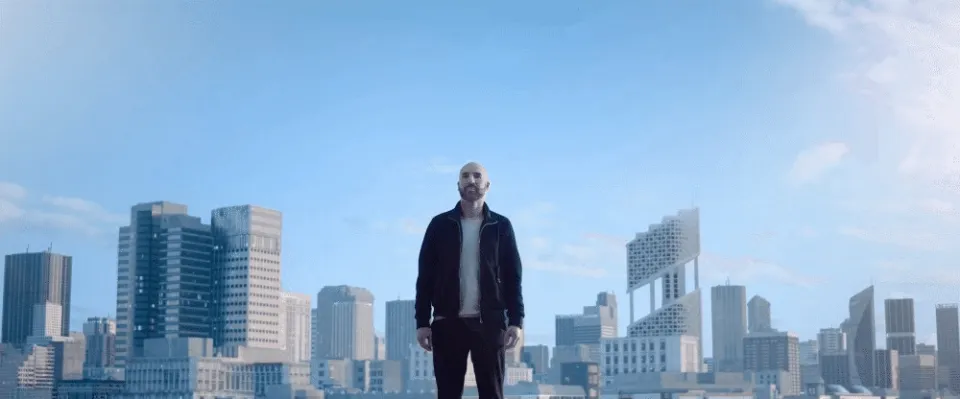 Mann in dunkler Jacke steht vor Stadtpanorama mit Hochhäusern unter blauem Himmel.