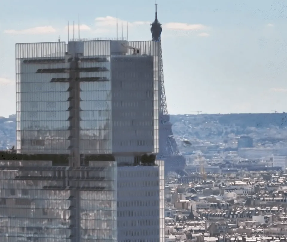 Modernes Hochhaus mit Antennenmasten im Vordergrund, dahinter der Eiffelturm und die Pariser Stadtlandschaft unter bewölktem Himmel.